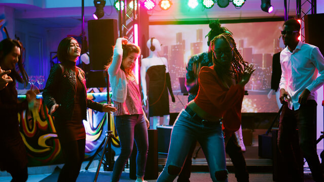 Funky people partying on dance floor enjoying electronic music party at nightclub. Group of happy friends dancing and partying together, having fun at social gathering. Handheld shot.