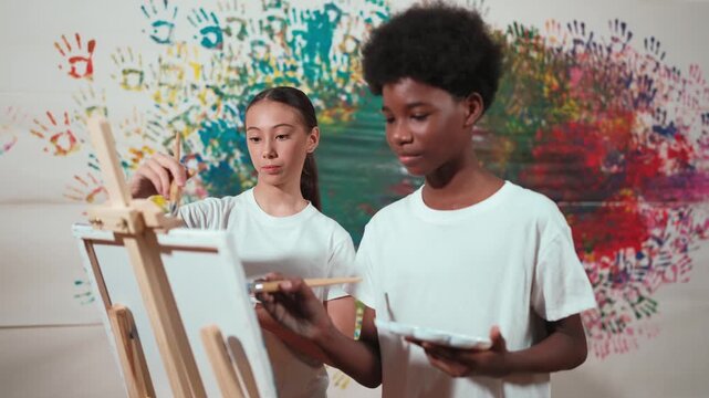Diverse happy children paint canvas together with colorful stained wall. Smart multicultural student drawing together in art lesson while wearing white shirt. Creative activity concept. Edification