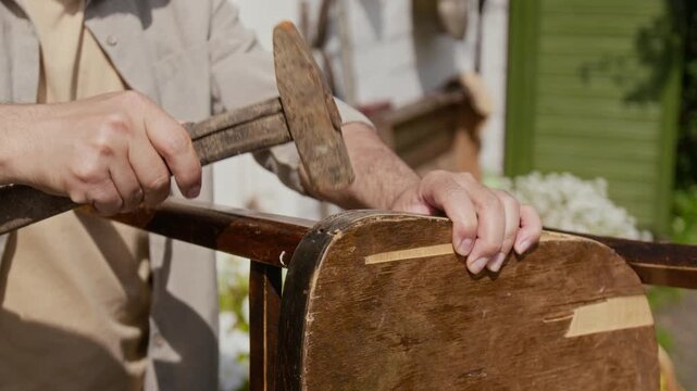Medium close-up of hands and midsection of thrifty country man restoring old beaten chair in garden, gluing together broken seat, fixing with hammer, displaying practicality and sustainable lifestyle