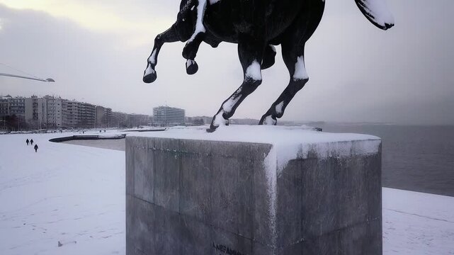 Drone ascending over the snow-covered Alexander the Great statue, revealing the winter coastline and Thermaikos Gulf in Thessaloniki.