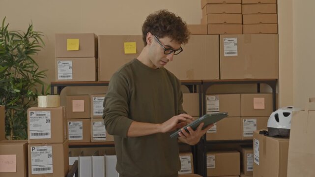 Man holding tablet and checking stacked packages while tapping labels in a shipping building; diligence efficiency.