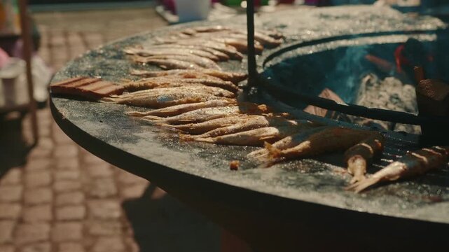 Fresh small fish grilling on a hot metal disc over an open fire. Street food cooking process outdoors. Traditional barbecue preparation for meal, picnic, summer festival or catering.