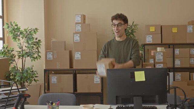 Man holding parcel at building desk near computer monitor and shelves, smiling while inspecting shipping label; small business joy.