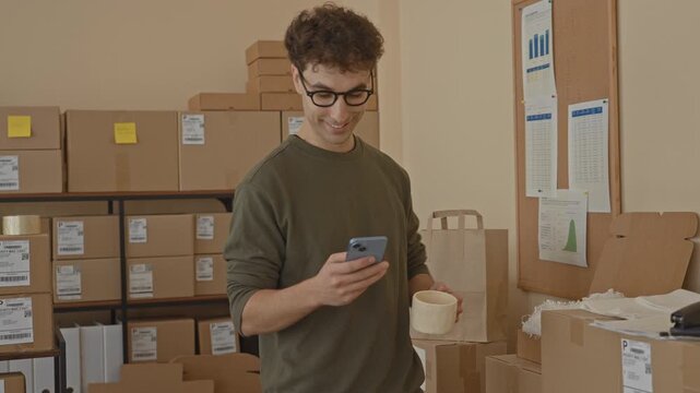 Man holds smartphone and coffee mug while checking parcels in a packing area inside a building; focused small business.