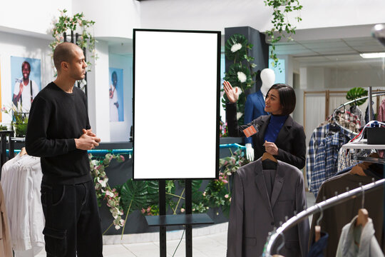 Boutique assistant guiding customer through blank interactive whiteboard mockup showcasing clothing collection. Store asian woman employee showing arab man client white ads display