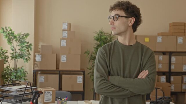 Man with arms crossed beside stacked cardboard boxes in a packing building; small business confidence.