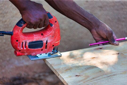 African Carpenter Using Electric Jigsaw to Cut Plywood Board Outdoors cut through a light wood