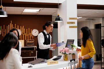 Fototapeta premium Hotel guest receiving registration form from helpful asian male concierge. Relaxed asian middle aged woman on vacation making necessary preparation for check in at modern resort