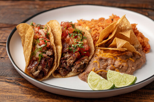 Two Carne Asada tacos on a white plate with rice, beans, cheese, chips and limes on a wooden table 
