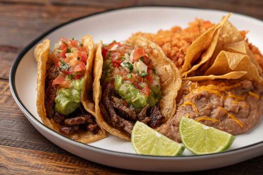 Two soft carne asada tacos on corn tortillas with guacamole, tomato, lettuce with beans, chips, and rice on a white plate, wooden surface. 
