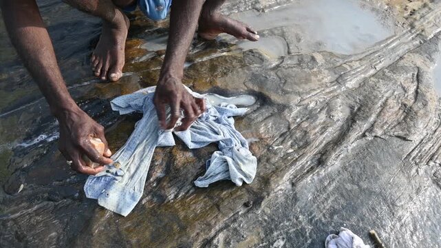 A man is washing clothes by hand on a stone at the riverbank. He uses soap to rub the fabric against the surface of the stone. How to wash clothes in the traditional way.
