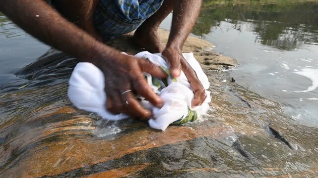 A man is washing clothes by hand on a stone at the riverbank. He uses soap to rub the fabric against the surface of the stone. How to wash clothes in the traditional way.