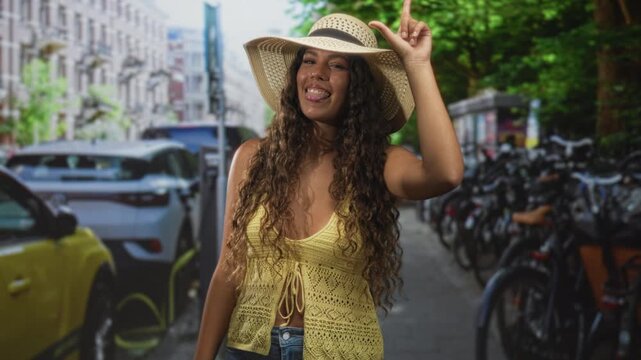 Woman tipping straw hat with index finger, yellow crochet top and midriff exposed on a street lined with bicycles and parked cars; summer fun playful.
