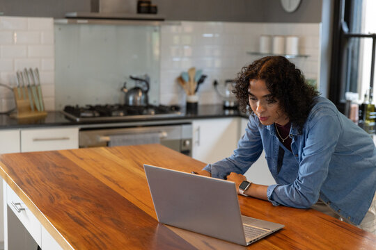 Non-binary adult leaning on wooden island, typing on laptop in kitchen with smartwatch, copy space