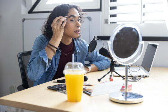 Non-binary mid-20s grooming eyebrows at home desk with mirror and mic using tweezers in denim shirt