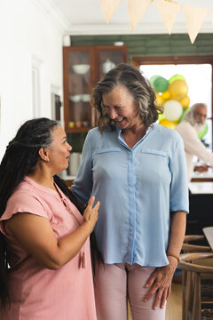 Diverse females in kitchen, left gesturing in pink top, right smiling in blue blouse near balloons