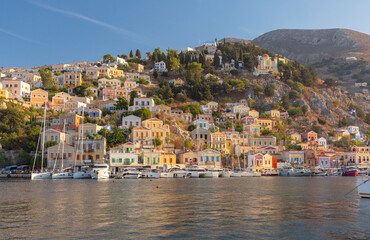 Fototapeta premium View of sailboats in the harbor of Symi, Greece, with colorful hillside houses, calm water and the church rising above the waterfront.