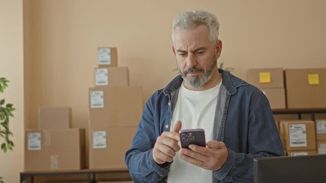 Man tapping smartphone screen while checking parcel labels amid stacked delivery boxes and a packing table in a building; concern logistics.