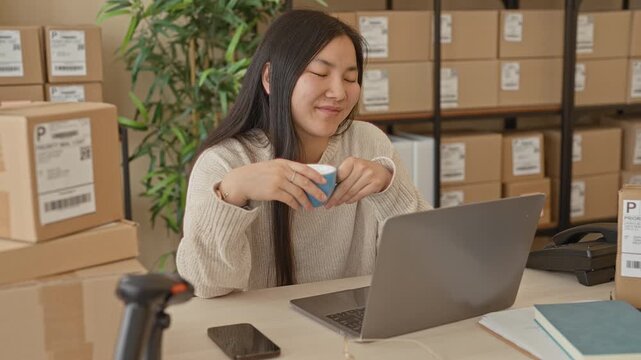 Woman holding coffee cup at laptop in a small business packing area inside building; calm productivity.