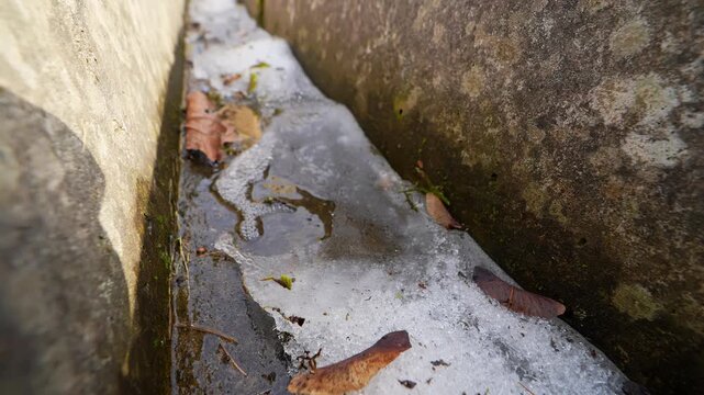 Thin melting snow and ice trapped in a narrow stone drain, with wet walls and fallen leaves showing seasonal thaw, runoff, and cold-weather surface texture.
