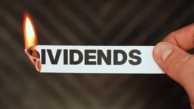Burning &ldquo;dividends&rdquo; label held by hand, symbolizing dividend cuts, suspended payouts, reduced passive income, and pressure on yield-focused investors.