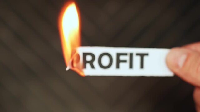 Burning &ldquo;profit&rdquo; label held by hand, symbolizing shrinking margins, business losses, earnings decline, and financial pressure on companies or investments.