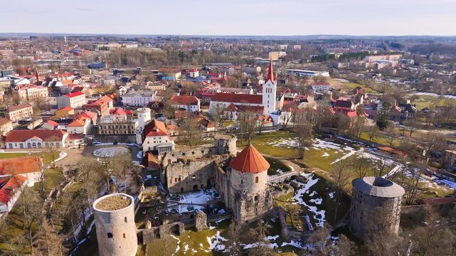 Aerial panorama of medieval castle ruins and a church-centered townscape, showing layered history, seasonal parkland, and compact urban character in soft daylight.