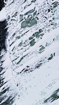Overhead close-up of a bootprint pressed into snow streaks on dark frozen glaze, revealing cracked patterns and a greenish tint&mdash;abstract winter hazard background, vertical.