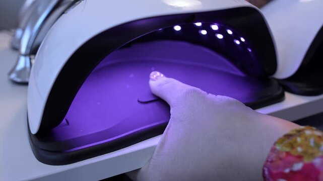 Close-up of a glitter thumbnail curing inside a UV LED nail lamp, highlighting gel polish hardening, violet salon light, and modern manicure technology.