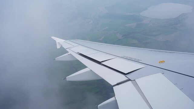Passenger window view of an airplane wing descending through cloud cover above countryside and a lake, capturing a moody in-flight landing approach.