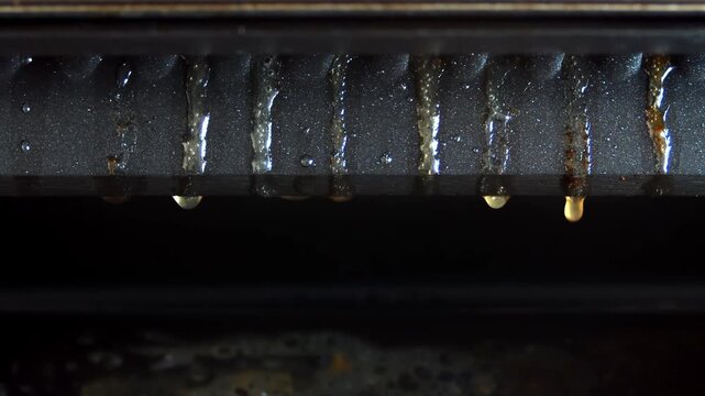 Close-up of grill tray surface as fat droplets slowly drip along textured ridges, shiny oily texture highlighted under direct light