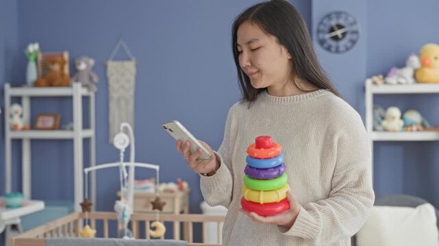 Woman holds smartphone and colorful stacking rings beside a crib in a nursery building; caregiving patience tired.