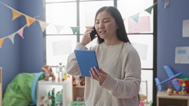 Woman holding tablet and smartphone to ear while looking up in a toy filled playroom inside a building; calm concentration.