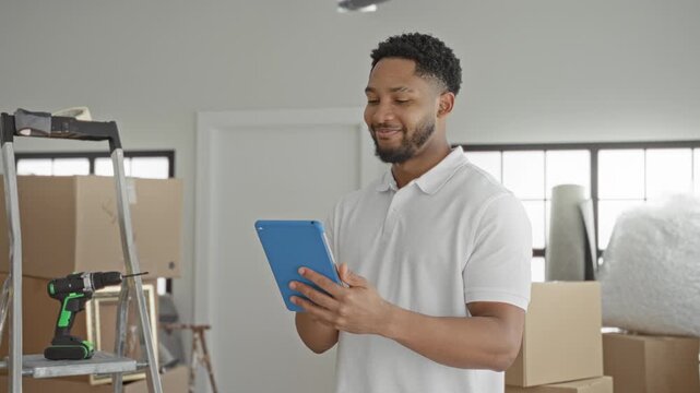 Man holding tablet and tapping screen while surrounded by stacked moving boxes, ladder, drill and wrapped furniture inside building; planning organization focused.