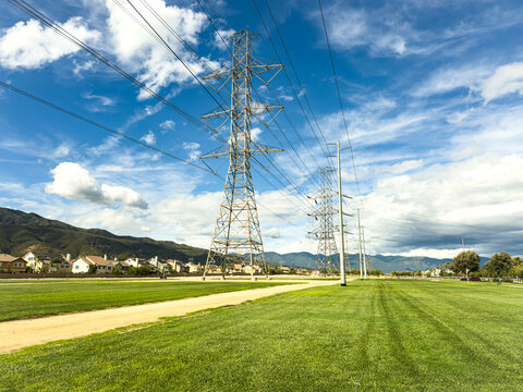 Tall metal power lines stretch across a green field under a bright blue sky with scattered clouds distant mountains and houses line the horizon creating a peaceful landscape