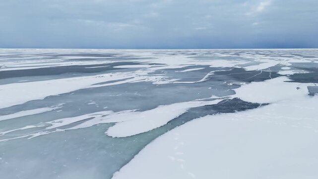 Drone shot gliding past a jagged ice pile into a vast frozen expanse with snow patches and dark ice lanes under a cold blue overcast sky.