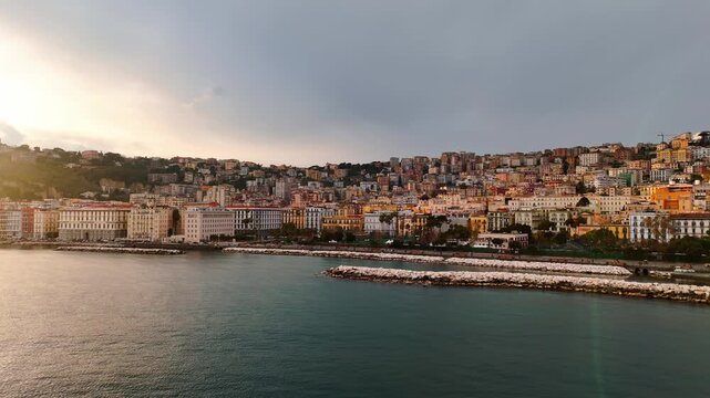 Aerial view of Naples, Italy, showing the bayfront boulevard, white breakwater, and tightly packed hillside neighborhoods glowing in soft evening light.