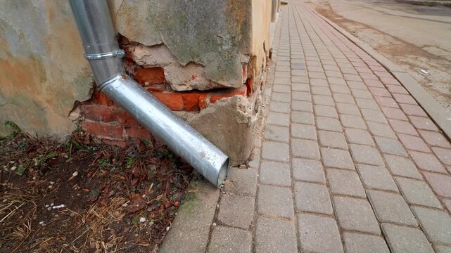 Metal downspout ends at a sidewalk beside crumbling plaster and exposed brick&mdash;poor drainage and weather damage on an old building exterior.