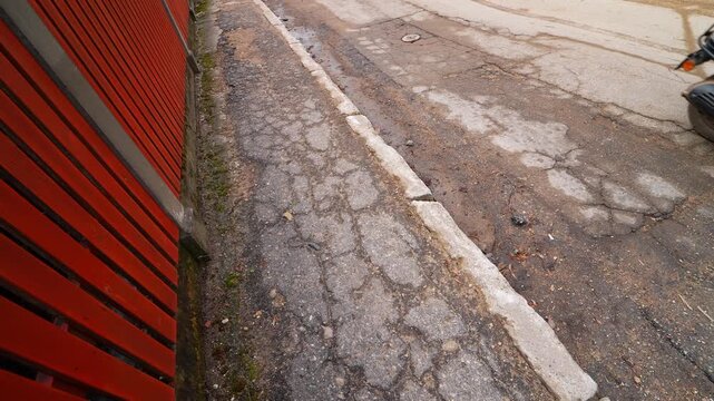 Cracked curbside asphalt and patched lane beside a red fence, showing gritty post-winter wear as a scooter passes&mdash;urban infrastructure stress and thaw damage.