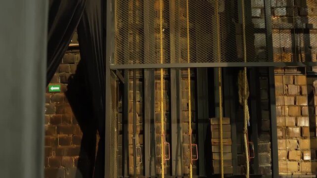 Wide backstage view of a theater fly system with counterweight arbors, ropes, steel framing, and brick walls, revealing the hidden mechanics behind stage scenery movement.