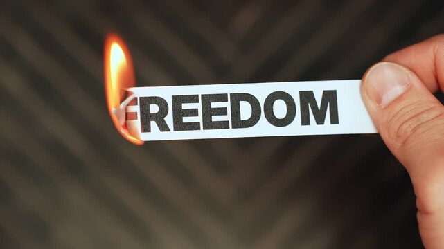Burning &ldquo;freedom&rdquo; label held by hand, symbolizing restriction, censorship, lost rights, control, and the threat of reduced personal liberty or independence.