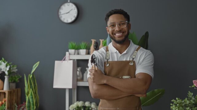 Man florist smiles with arms crossed holding pruning shears, bare forearms visible, wearing apron and glasses in a flower shop filled with potted plants and vases; welcoming warmth.