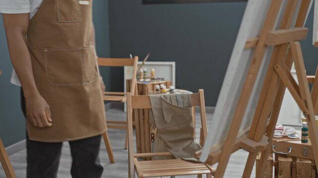 Man painter touching chin at easel in studio, seated on stool wearing apron and glasses; quiet contemplation.