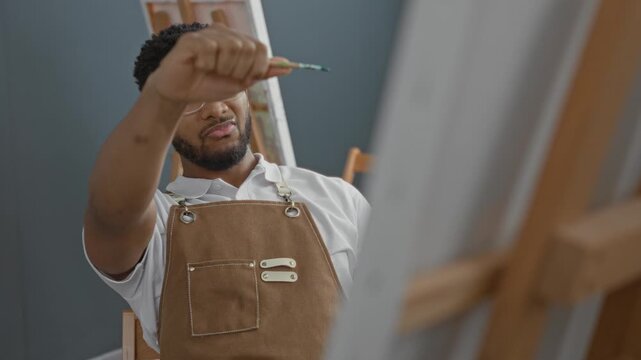 Man painter in studio holds brush to canvas wearing apron and clear glasses, painting with steady hand and intent expression; quiet concentration.