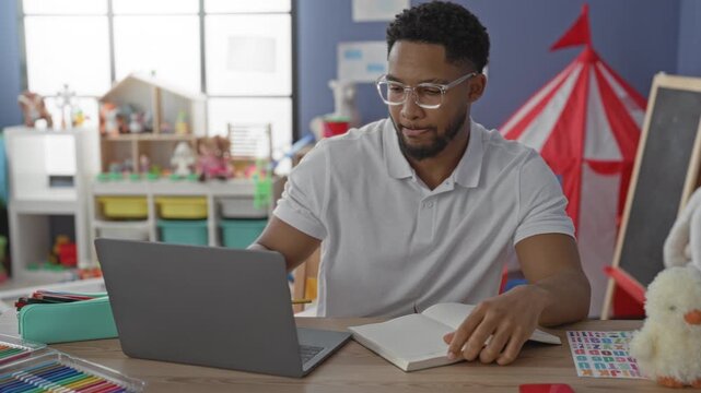 Man writing with pencil at laptop and notebook in colorful preschool classroom with toys, chalkboard and play tent; focused planning.