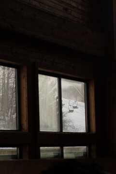 View from inside a rustic cabin looking through a window toward a snowy mountain slope with an empty ski lift. The scene combines a warm indoor atmosphere with a cold outdoor winter landscape