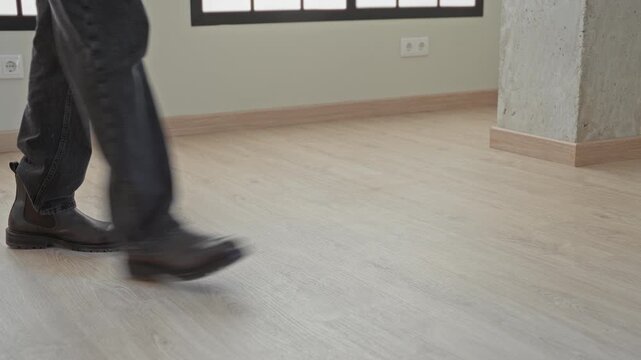 Man walking in brown leather boots and dark jeans across light wood floor near concrete pillar and window in studio; quiet solitude.