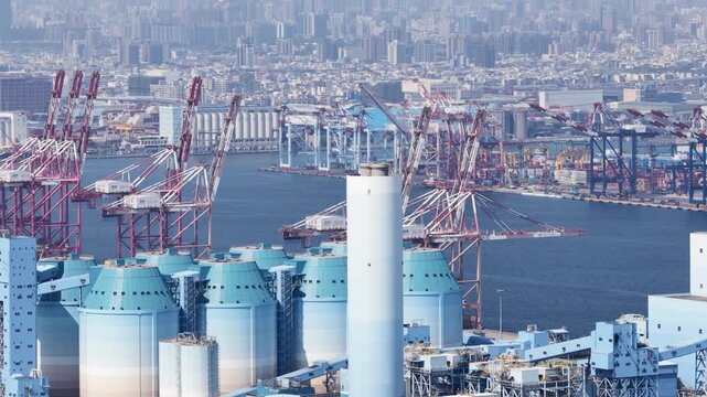 Massive industrial port featuring large silos and numerous gantry cranes for loading and unloading cargo ships, with a sprawling urban cityscape visible in the hazy background on a sunny day