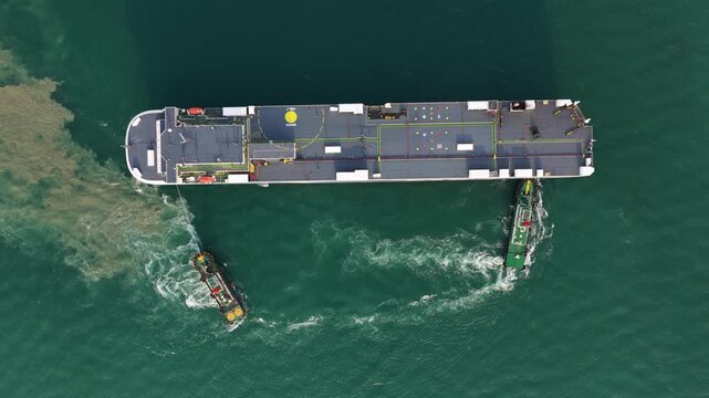Aerial top down drone video of a tug boat escorting a large ship out of port, showing harbor operations, maritime transport, vessel guidance, and commercial shipping movement.
