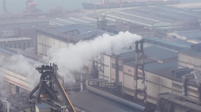 Aerial view of a large steel factory with chimneys emitting thick white smoke, contributing to air pollution and global warming in a heavily industrialized area with a nearby port